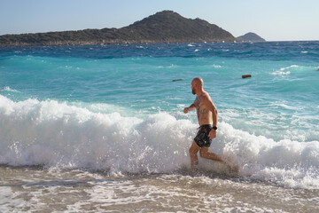 Young tattooed sporty man on the beach on a hot summer sunny day, playing with waves, jumping, having fun