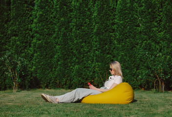 Blonde in sunglasses and headphones with mobile phone sitting in yellow bean bag in a garden