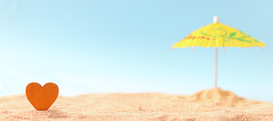 Close up look on orange wooden heart stuck in the sand with open parasol in background on the empty...