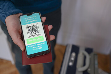 Vaccine passport, close up picture of male hand holding smartphone and showing International certificate of vaccination and suitcase.