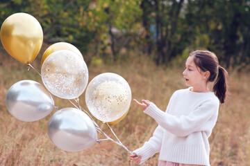 Surprised happy teenage girl in a white sweater holding a lot of gel balloons that fly away from the wind