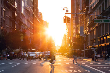 Busy street scene in New York City with crowds of people walking across an intersection on 5th Avenue with the light of summer sunset shining in the background