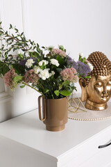 Stylish ceramic vase with beautiful flowers and golden Buddha sculpture on chest of drawers near white wall