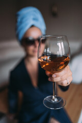 A girl at home drinks wine in sunglasses after a shower. Glass in focus. A blue towel is wrapped around the girl's head. There's white wine in the glass. The girl holds out a glass to the camera