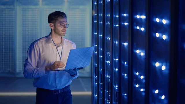 Male Server Engineer in Data Center. IT engineer inspecting a secure server cabinet using modern technology laptop coworking in data center.