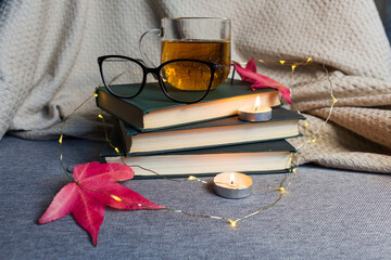 a cup of tea stands on the books next to glasses, candles and autumn leaves on a blanket with lights
