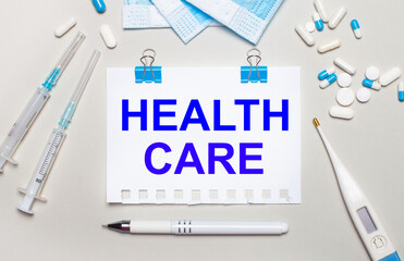 Fototapeta premium On a light gray background, blue medical masks, syringes, an electronic thermometer, pills, a pen and a notebook with the inscription HEALTH CARE. Medical concept