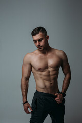 A man with a naked torso in the studio. The man leaned back a little, holding on to his belt with one hand. The muscles of the abs are clearly visible, the gaze is directed downwards