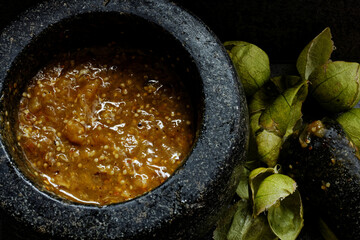 Roasted tomatillo salsa with toasted chilies de arbol in molcajete and tomatillo husks - obrazy, fototapety, plakaty