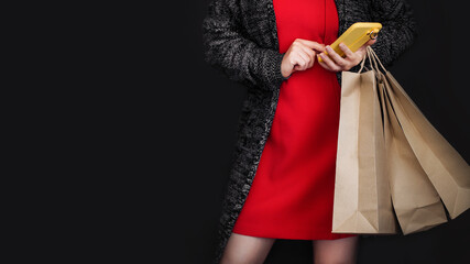 A woman uses her phone and holds Black Friday shopping bags while standing against a black background.