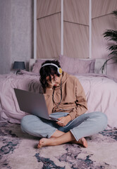 a frustrated teenage girl sits near the bed and learns remotely via a laptop and headphones