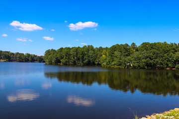 Obraz na płótnie Canvas a gorgeous shot of vast still blue lake water surrounded by lush green and autumn colored trees with blue sky and clouds at Lake Peachtree in Peachtree City Georgia