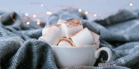 Adding cacao powder on top of hot chocolate with marshmallows in white cup on sweater background. Cozy christmas lights.