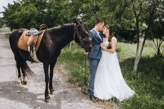 A young groom in a suit and a beautiful bride in a white long dress hug, smile and kiss in a village outdoors, walking with a black horse along a country road. Wedding photography, portrait.