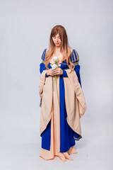 Fototapeta premium An attractive woman with long hair in a medieval, fantasy, blue and beige dress with long, large sleeves, posing with white roses in her hands, isolated on a white background.