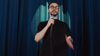 Young emotional guy comedian wearing eyeglasses performing his stand up show, interacting with audience in comedy club, moving on stage
