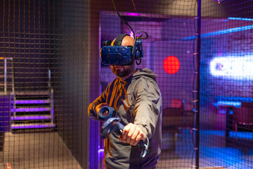 young bearded man in virtual reality helmet and with controller in his hands plays online game in VR club, game simulates behavior in fictional world, gadgets and virtual reality addiction