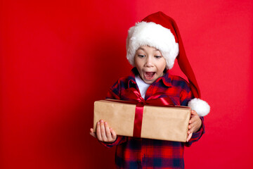 Happy holiday and Merry Christmas. Portrait of a funny surprised boy in a cap with gifts in his hands on a red background. A place for text.