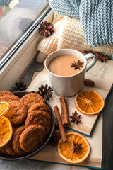 A large cup of coffee and a book near the window, several knitted blankets and slices of dried oranges remind of autumn, a cozy time and home holiday. Flat lay