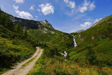 steep dirt road to a long white waterfall in a green mountain landscape
