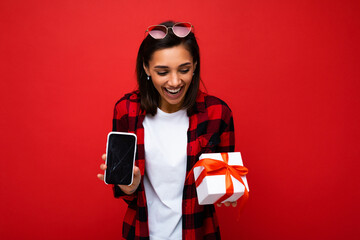 Attractive happy young brunette woman isolated over red background wall wearing white casual t-shirt and red and black shirt holding white gift box with red ribbon and mobile phone with empty screen