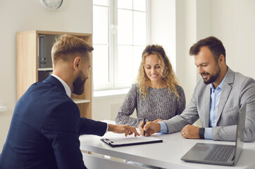 Young married couple decide to buy or rent house. Happy husband and wife sitting at office table and signing contract agreement given by professional realtor or real estate agent - obrazy, fototapety, plakaty