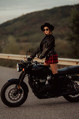 Obraz na płótnie Canvas Stylish motorcyclist woman in mini skirt, leather jacket stands on vintage-styled motorcycle. Attractive alluring driver in jackboots on highway. Trip, speed, style, feminism concept.