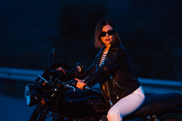 Fototapeta na wymiar Hipster stylish motorcyclist woman sitting on vintage-styled motorcycle. Young female driver night on roadway. Trip, freedom, classic motorbike concept.