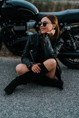 Fototapeta na wymiar Motorcyclist woman in mini skirt sitting on the asphalt near her retro-styled motorcycle. Sexy female driver in jackboots and sunglasses on roadway. Trip, speed, freedom concept.