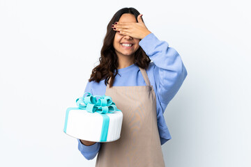 Pastry chef holding a big cake over isolated white background covering eyes by hands and smiling