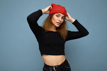 Young pretty positive brunette woman curly with sincere emotions wearing stylish black crop top and red hat isolated on blue background with copy space