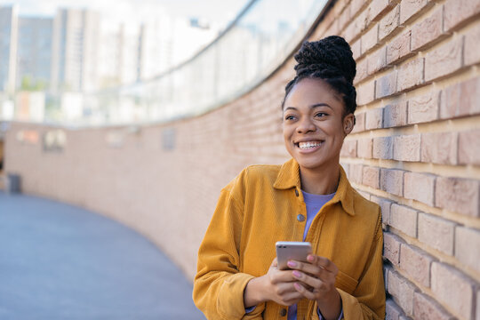 Beautiful smiling African American woman using mobile app shopping online with sales standing near wall. Smiling hipster female wearing stylish yellow shirt holding smart phone text messaging 