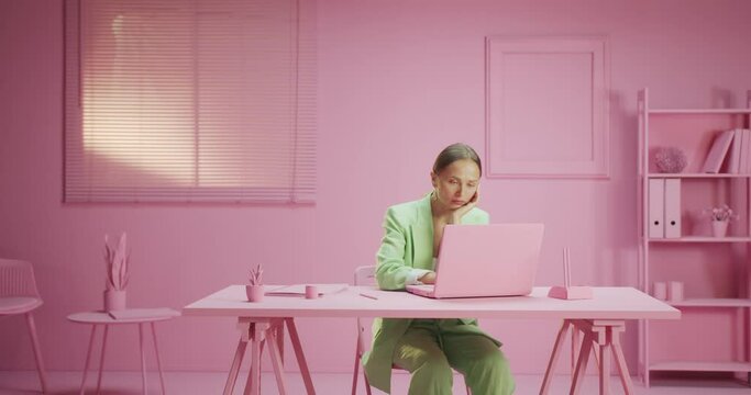 Horizontal moving shot of sad and tired young female worker at pink workplace indoors, exhausted beautidul woman wearing light green costume sitting staring at computer depressed and unhappy at work
