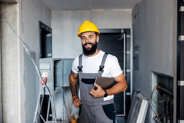A happy construction worker with a helmet is standing in an unfinished building and holding a...