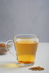 Helba golden tea drink made of fenugreek plant served in glass cup with seeds on white wooden background against gray wall at kitchen used as food ingredient. Vertical image with copy space