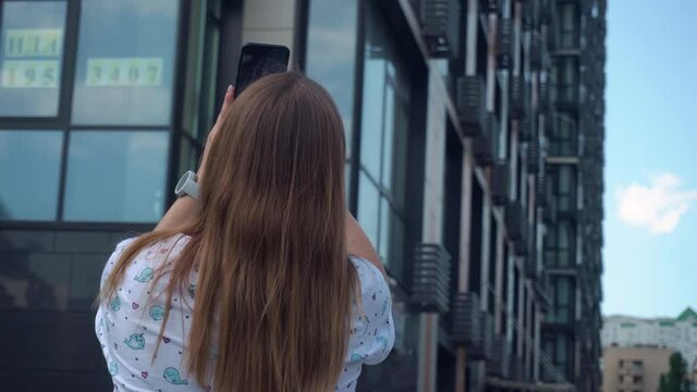 Beautiful girl with long blond hair in sunglasses and smart watch on her hands in a white T-shirt with whale makes video on her phone. against the background of a tall new building. summer, clear day