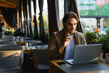 Stylish freelancer having video call on laptop on terrace of cafe in evening 