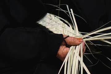 Hands of elderly arab woman artisan in traditional black dress close up weaving manually basket from dried organic palm leaves. Arabian traditional crafts.  - obrazy, fototapety, plakaty