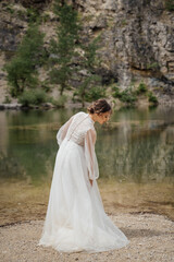 view from back. bride in a white dress stands on the shore of a mountain lake.