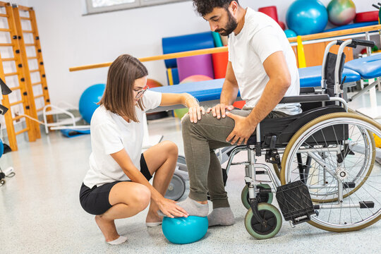 Pphysiotherapist and male patient seated in a wheelchair during rehabilitation treatment - press and release a foot on a small exercise ball