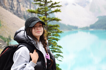 Fototapeta na wymiar Young girl with backpack standing in front of turquoise lake and mountains smiling looking at camera