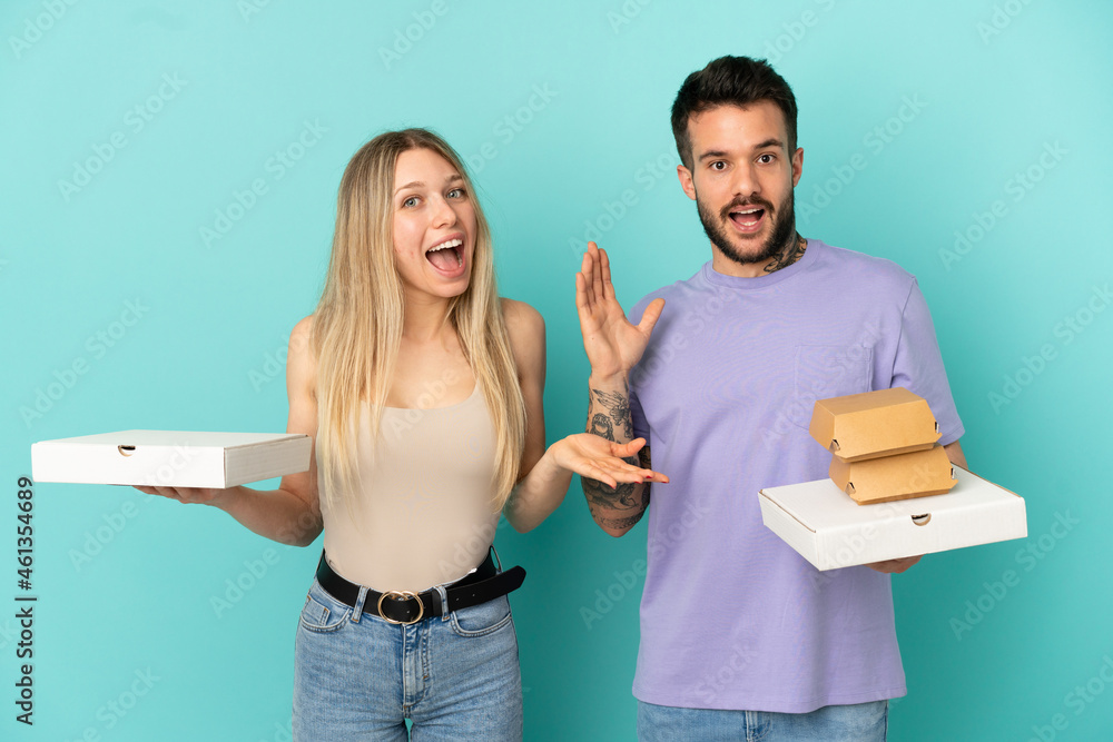 Wall mural Couple holding pizzas and burgers over isolated blue background with surprise and shocked facial expression