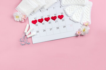 Monthly calendar with marked menstruation days and various types of feminine sanitary pads and tampons decorated with pink flowers. Female menstrual cycle concept. Copy space