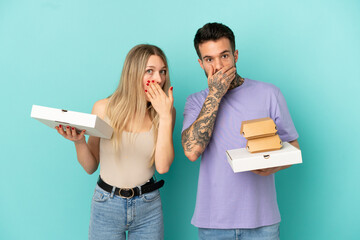 Couple holding pizzas and burgers over isolated blue background covering mouth with hands for saying something inappropriate - obrazy, fototapety, plakaty