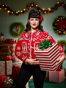 Portrait of woman with a retro hair style, in ugly Christmas sweater holding present looking at camera.