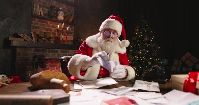 Santa Claus working at night, opening letters from kids close-up. St Nicholas reading children wishes for preparing presents. Late work in evening on Christmas Eve. Green tree on background.