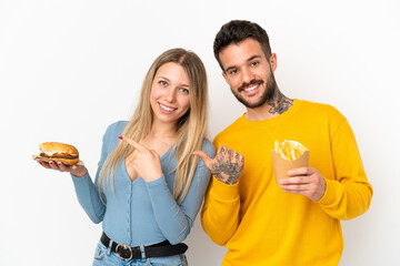 Couple holding hamburger and fried chips over isolated white background pointing finger to the side...