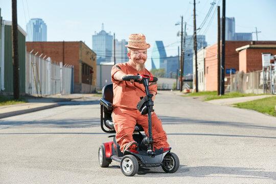 Portrait of Caucasian male adult little person on a mobility scooter parked in middle of street with Dallas Texas skyline in background.