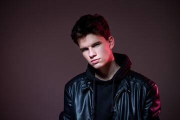 Attractive young guy posing in studio. Young caucasian guy model in black clothes posing in the studio on a dark burgundy background with red backlight. Man in a leather jacket.