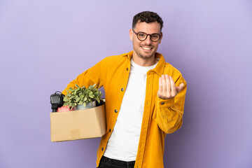Young caucasian man making a move while picking up a box full of things isolated on purple...